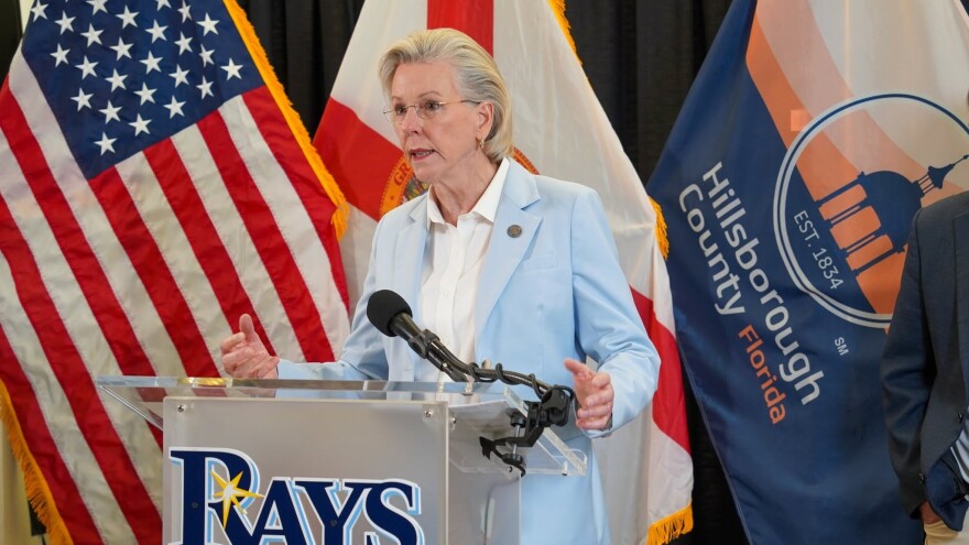 older women with short blond hair wearing a light blue pants suit with blazer. is speaking at a podium that has the Tampa Bay Rays logo. Flags of the US, Florida and Hillsborough County are behind her