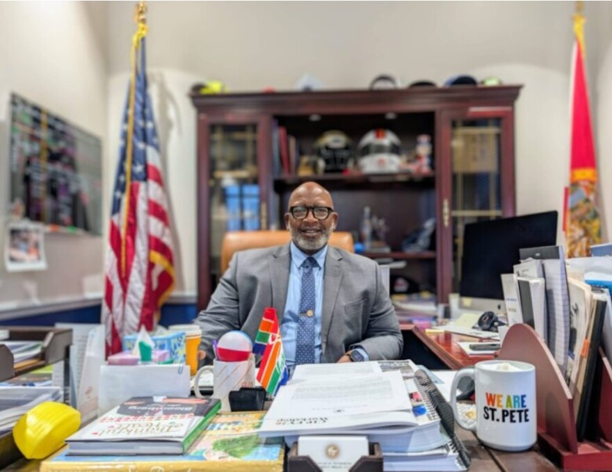 Man in a gray suit and blue tie sitting behind a desk with books and a We Are St. Pete mug in the foreground