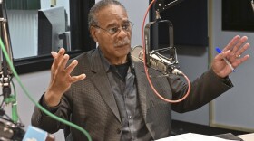 A man sitting inside a radio studio sits behind a microphone. He is talking and gesturing with both hands.