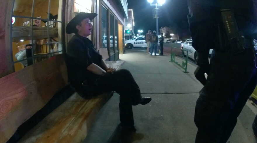 A woman in a cowboy hat sits on a bench and talks to a cop. Cop cars are in the background.