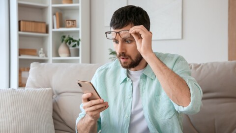  a shocked young man with eyeglasses stares at his mobile phone as he sits on the sofa