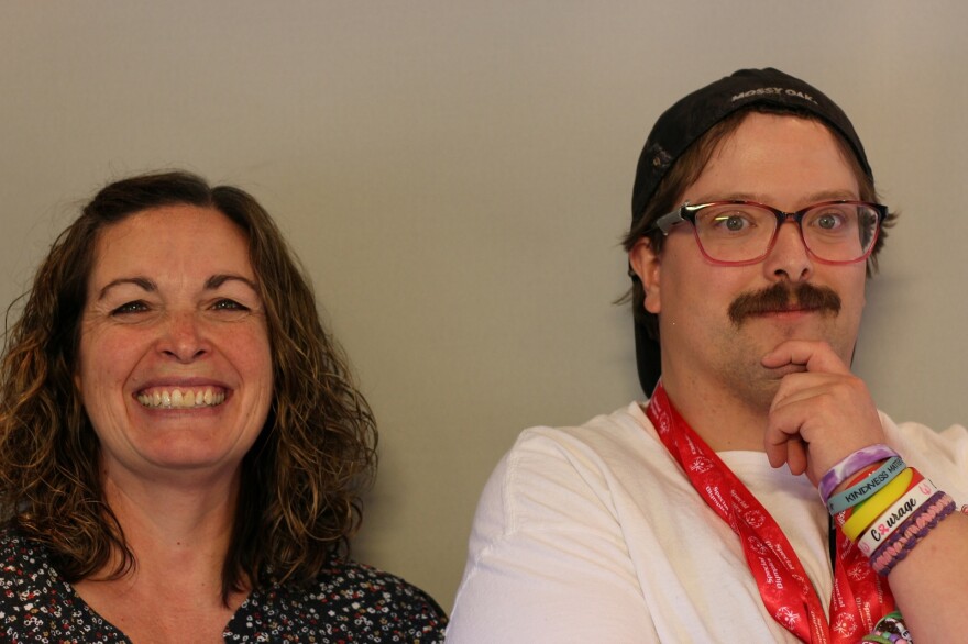 Karen Willmott and Steven Mongtomery at their St. George StoryCorps appointment in May 2025.  Karen Willmott faces the camera fully and smiles widely, teeth showing, and it makes her eyes small.  She has shoulder length, brown hair that is worn parted on one side, clipped back with a barrette on the other, and wavy.  She has on a dark blue top with a small floral print.  Steven Montgomery poses with Karen to his right. He is turned slightly away from her, with one hand on his chin.  He wears a backward baseball cap and glasses, and he has a full moustache.  On his wrist, he wears a number of plastic bracelets.  Around his neck are orange ribbons that hold his sporting medals.  He has on a white tee shirt.