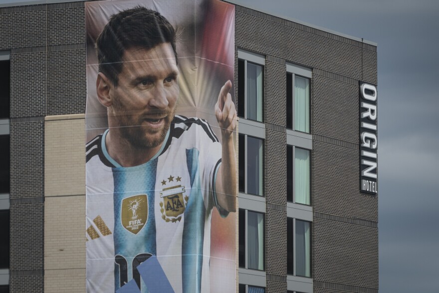 A large brick building is partially shone. A large, two-story poster hangs on one corner of an Argentinian soccer player pointing his finger.
