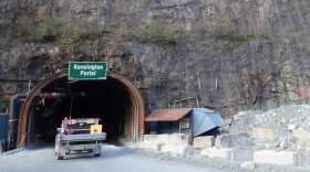 A truck enters a tunnel in a granite mountain. There is a green sign that says "Kensington Portal" above the tunnel and a blueish vent on the right side next to the vehicle.