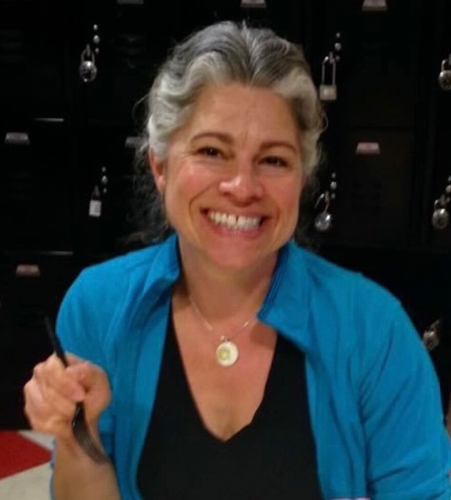 A woman with grey hair wearing a blue jacket, holding a fork and smiling at the camera