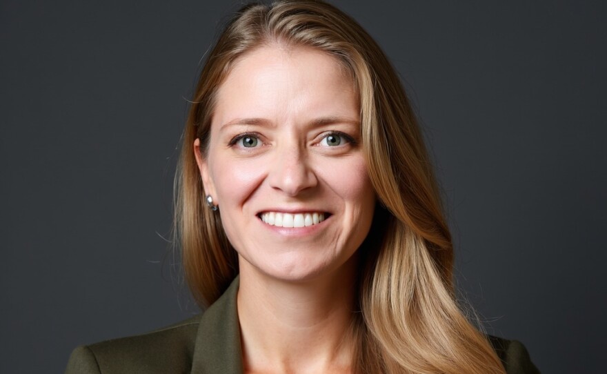 A smiling woman with long blonde hair wears a white shirt under an army green blazer.