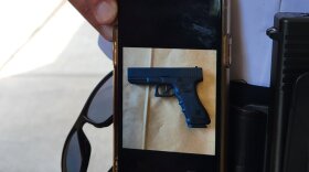 A white police captain holds a iPhone at his hip, between his gun and his sunglasses. The iPhone is showing a horizontal photo of a black Glock-style BB handgun against a brown background. 