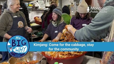 People gather in a cozy, bustling kitchen space at Winter Kissed Farm in Montana’s Bitterroot Valley, preparing kimchi during a traditional Korean kimjang event. In the center, Sohn Jung-a, one of the organizers, smiles while helping mix ingredients in a large metal bowl. Around her, other participants in winter clothing work together, handling napa cabbage coated in red spices. A large tub of kimchi sits in the foreground. A banner reads: "The Big Why – Kimjang: Come for the cabbage, stay for the community."