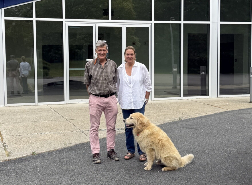 Man with gray hair in plaid shirt and pink pants standing beside a woman in white shirt and jeans and a golden dog. 