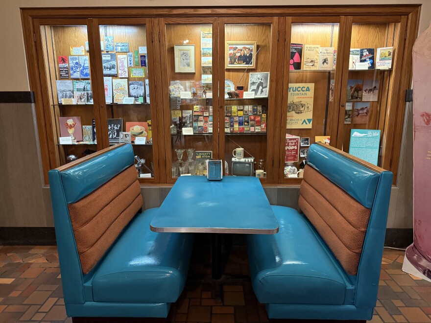 An old blue and brown diner booth sits inside a coffee shop, with a trophy case full of knick-knacks, papers and pictures behind it.