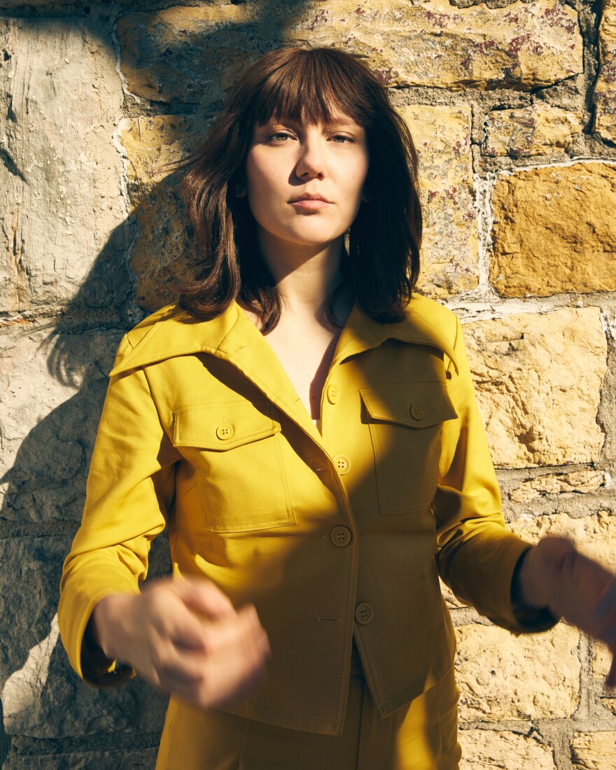 A woman looks at the camera while standing in front of a stone wall.