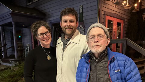 Three people stand on a sidewalk in the evening.