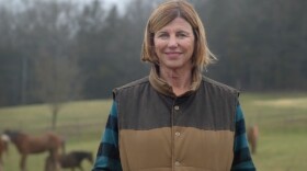 Woman wearing a cold-weather vest and long-sleeved shirt standing in a field with horses in the background.