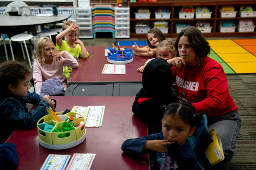 A woman sits at a low table talking to kindergarten students.