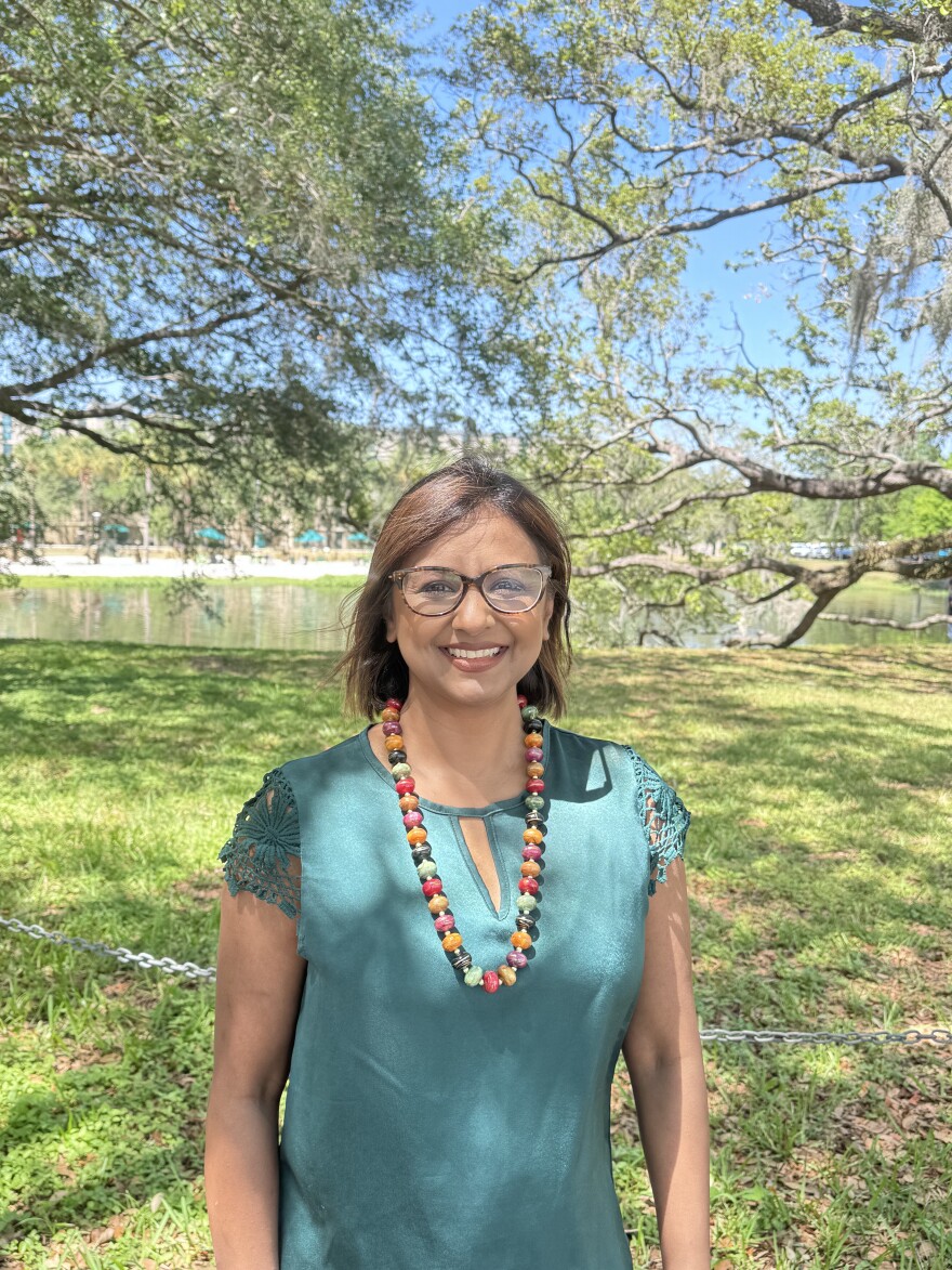 A woman stands in front a tree with a pond in the background. She's wearing a green shirt and a brown beaded necklace.