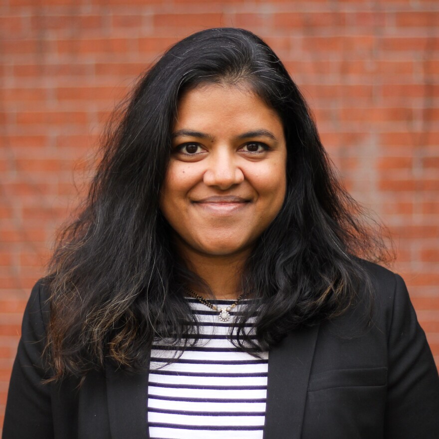 A professional headshot of a smiling woman with dark hair past her shoulders and golden brown skin.