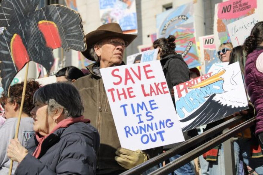 A man wearing a brown felt cowboy hat and brown zip up jacket looks to his left as he stands with other people on a staircase outside. He holds a sign that says "SAVE THE LAKE TIME IS RUNNING OUT."  About eight other people are seen in the photo and most of them are holding protest signs. The building's wall is in the background, but the signs and people are mostly covering it up.