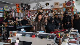 A woman with a large group of musicians sings while standing behind an office desk and in front of shelves full of albums and other music items.