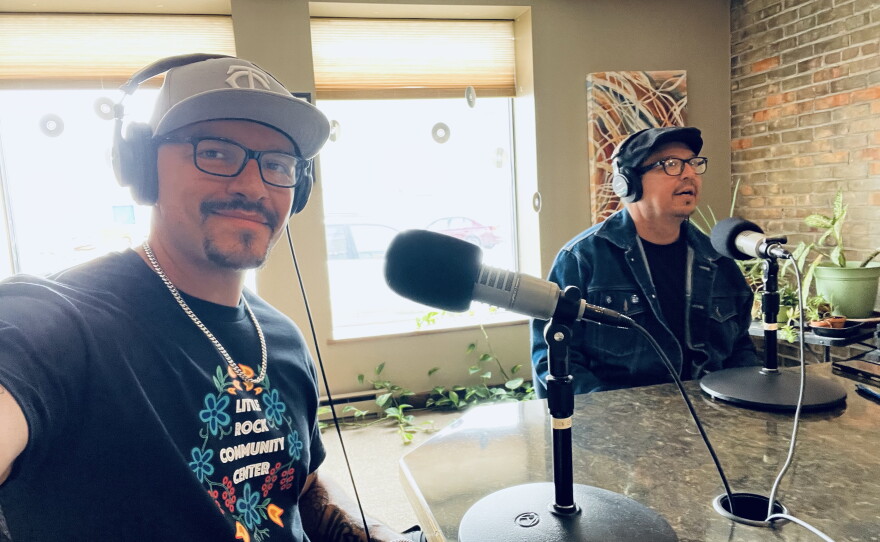 Two men in a radio studio sitting behind microphones. One is holding his arm out to take the selfie.
