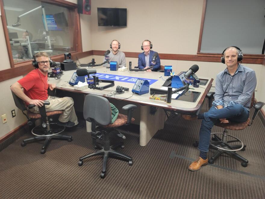 Four men wearing headphones sit at a table in a radio talk studio: a man front left has grey hair, a grey beard and is wearing glasses,  a red t-shirt and tan pants; a man front right has short dark hair and is wearing a grey button-down shirt, jeans and brown shoes; a man back left has short brown hair, a brown beard and is wearing a grey sweater; a man back right has short blonde hair and is wearing glasses, a navy blazer and a light blue button-down shirt.