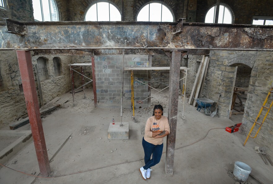 A woman leans against an upright, steel post that support a large steel beam inside a cavernous stone room. Other building materials and scaffolding are positioned in the background.