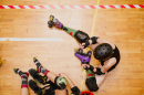 Two roller derby skaters collide while sitting on a wooden-floor rink, wearing helmets, knee pads, and roller skates.