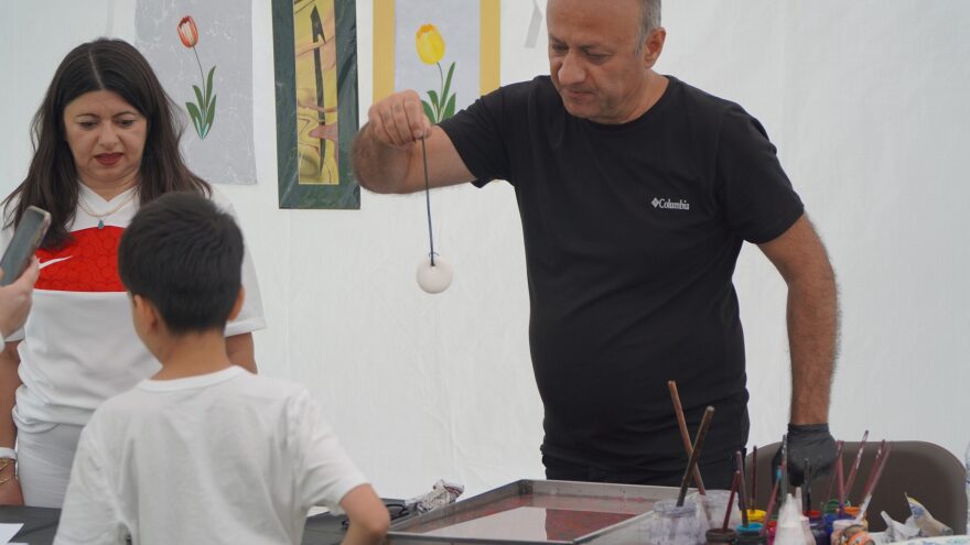 A man holds a stone above a tray full of pink water. A kid stands in front of it. A woman is next to the man, watching the process. There's paintbrushes and pigments next to the tray.