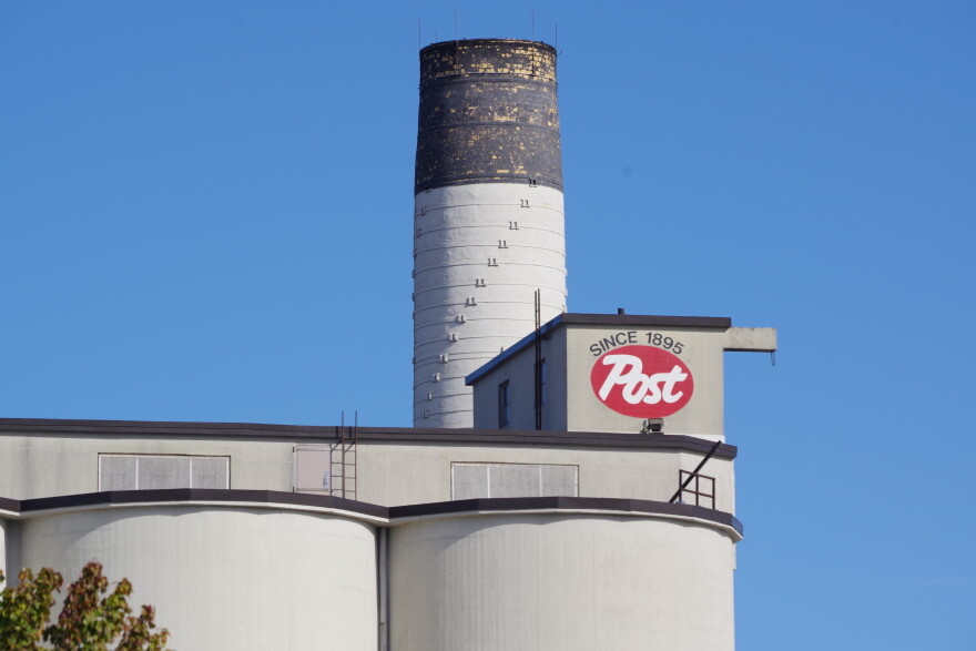 A red oval is painted on the side of a small structure at the top of large grain silos. "Post" is written inside the oval in white text, and "since 1895" is written above the logo. A chimney stands behind the structure, just a bit taller than the silos.