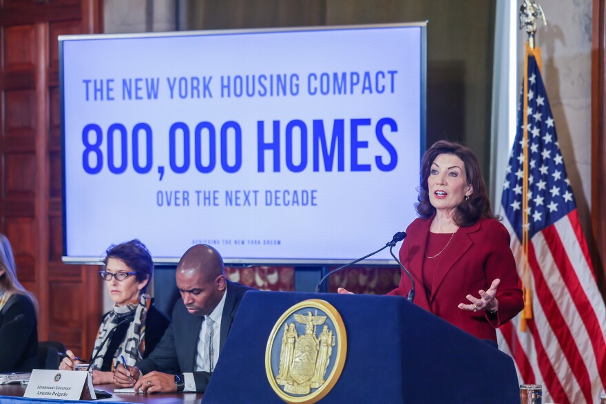 A woman in a red outfit stands behind a lectern next to a monitor that says "The New York Housing Compact: 800,000 homes over the next decade.