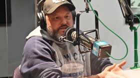 A man wearing a sweatshirt and baseball cap talks at a microphone inside a studio.