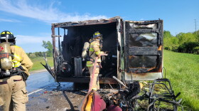Two firefighters stand with hoses, extinguishing  the remnants of a fire from near a charred van on rural higway.