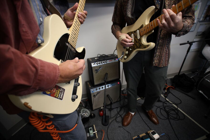 A close-up of two guitarists playing electric guitars with their amplifiers stacked on the floor.