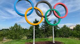 The blue, yellow, black, green, and red Olympic rings sit suspended in the air against a blue sky.