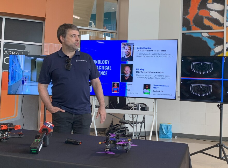 A man in a dark shirt and pants standing by table in large room. On the table in front of him are two drones.