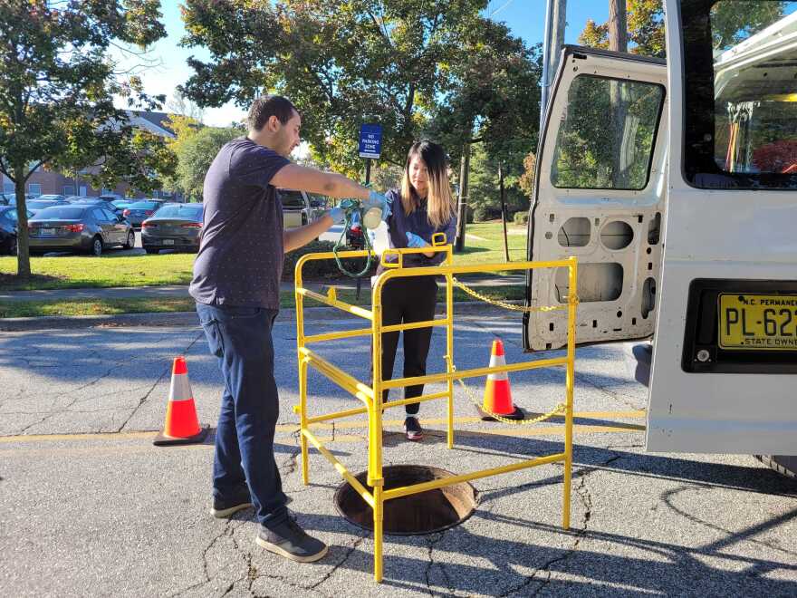 A man and woman are standing in front of a yellow barrier that is around a sewer hole. The man is pouring wastewater from the sewer into a collection bottle the woman is holding. 