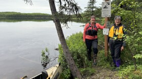 Two women in life jackets stand next to a canoe by a lake.