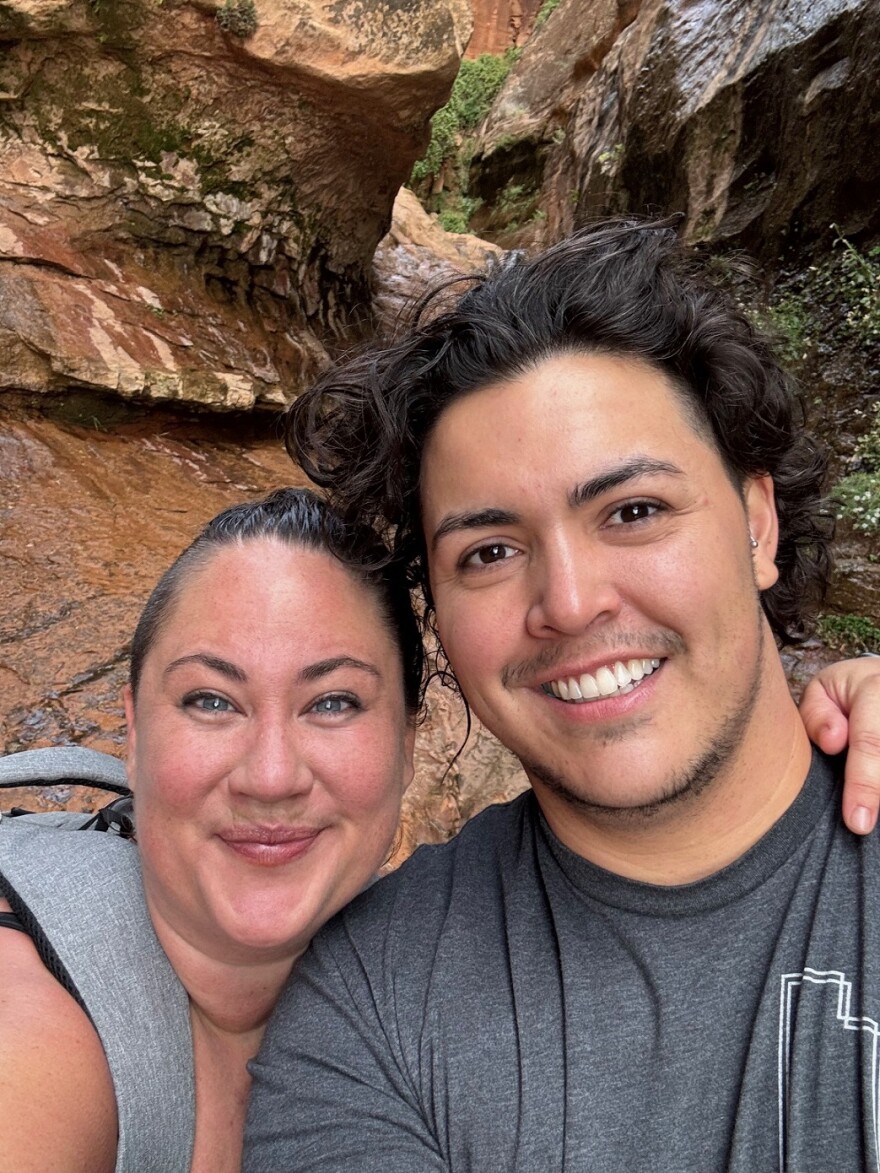 Morgan and Micah Barrick pose in a rocky hiking area.  Morgan has a grey padded hiking pack on over a strapped tank top.  She has one arm around Micah.  She smiles with a closed mouth and raised eyebrows. Her hair is pulled back in a pony tail.  Micah has dark hair and eyes and wears a thin, trim beard and mustache and a gray tee shirt with the outline of the state of Utah in the  upper corner.