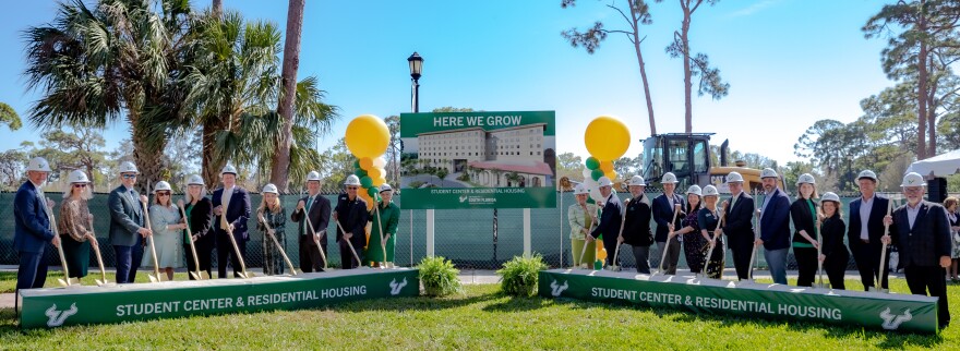 Group of people wearing hard hats and holding shovels stand outside. The shovels are set into two ceremonial sets of sandboxes with the words "student center and residential housing" on them. A sign with the words "here we grow" and the rendering of a building, along with balloons, are between them.