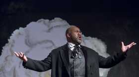 A singing man in an old-style black suit, hands outstretched and a grey cloud illustration in the background 