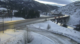 A wet road with a few cars on it winds through snowy hills and over a river.