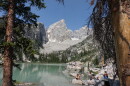 A rocky granite peak rises over an aquamarine lake. People sit and stand on rocks near the shore.