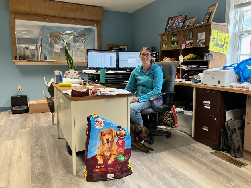 A person sitting at a desk, with her dog underfoot and two computer screens behind