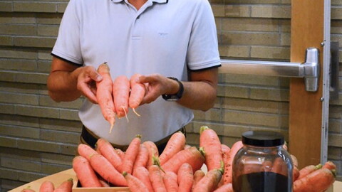 A man holds up three carrots, in front of a table of carrots and a jar of something that looks like coffee grinds.