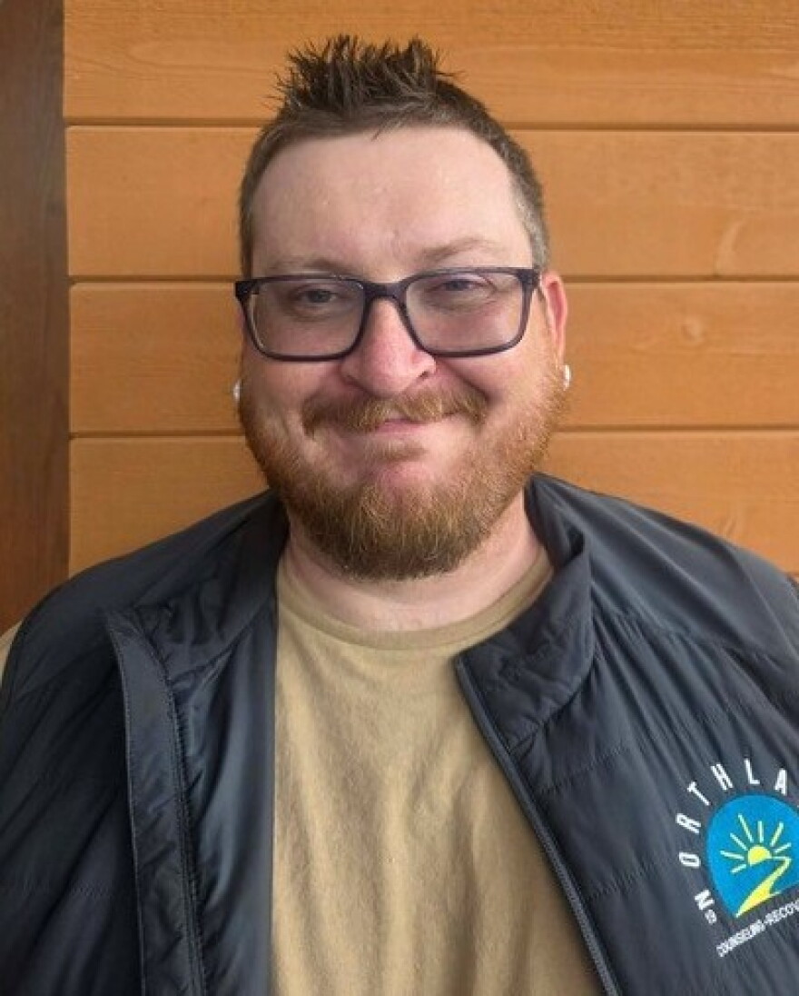 A person wearing a navy blue vest and yellow shirt underneath standing in front of a brown wall.