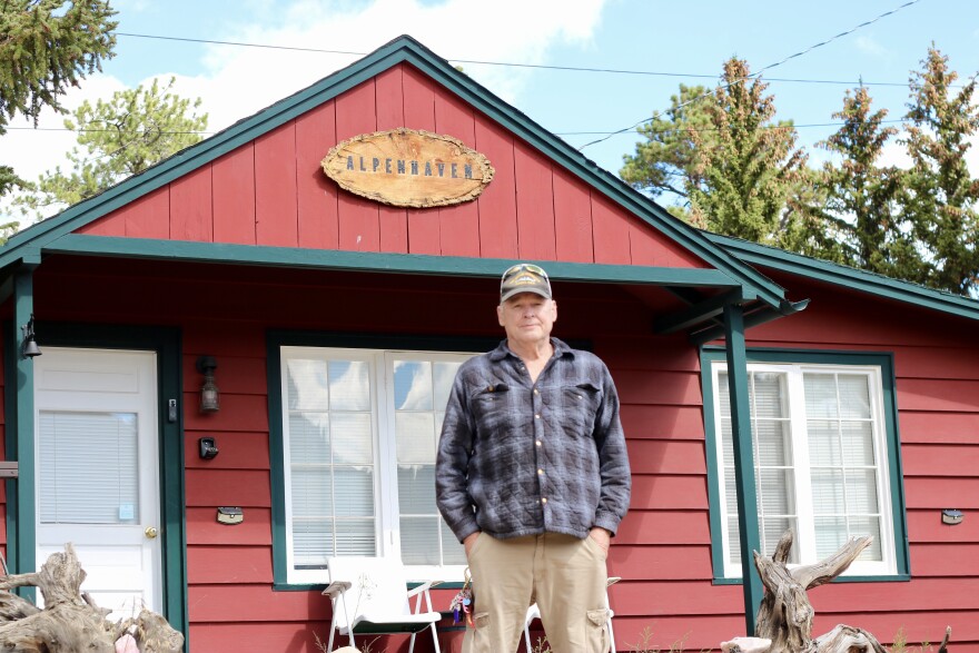 A man in a plaid shirt stands in front of a red cabin.