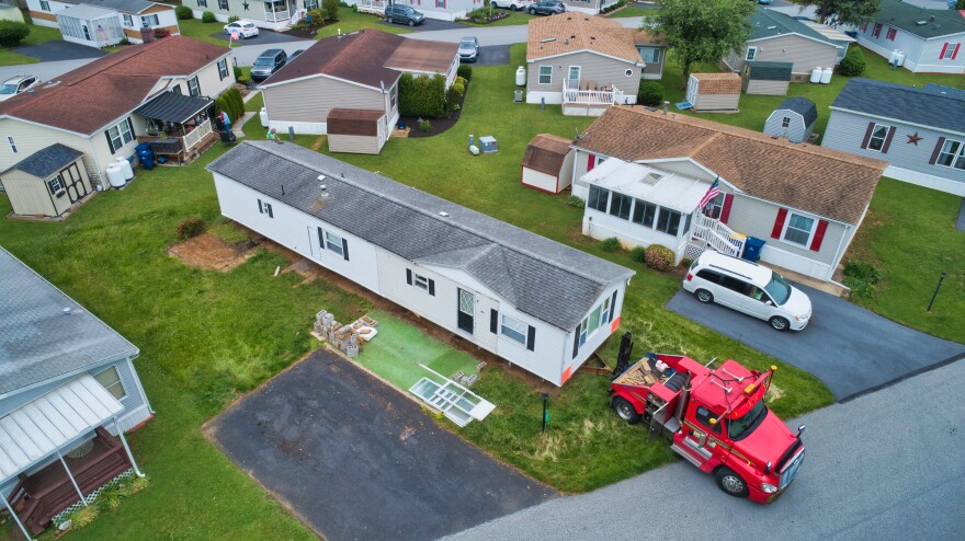 An aerial view shows a long white manufactured home beside a red tow truck in a neighborhood of similar homes. Soil is exposed along the home’s sides. Cinder blocks, window frames, and other materials sit near a driveway. Nearby homes have lawns, sheds, porches, and parked vehicles.
