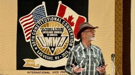 A white older man with a short mustache and beard  wearing a green and white flannel speaks towards a microphone with both of his hands turned up in front of him. In the background is a quilt that says 55th Constitutional Convention Las Vegas, NV 2015 on the wall. There's a backwards American flag to the left and a Canadian flag to the right on the quilt. At the bottom it says 1890 with a quilted ribbon leading towards the right side that the man is standing in front of.
