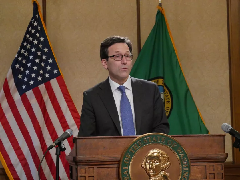 A man in glasses stands and speaks at a podium next to an American flag.