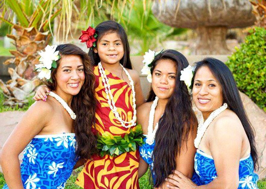 Four women dressed in traditional Polynesian dresses, leis, and flowers in their hair.