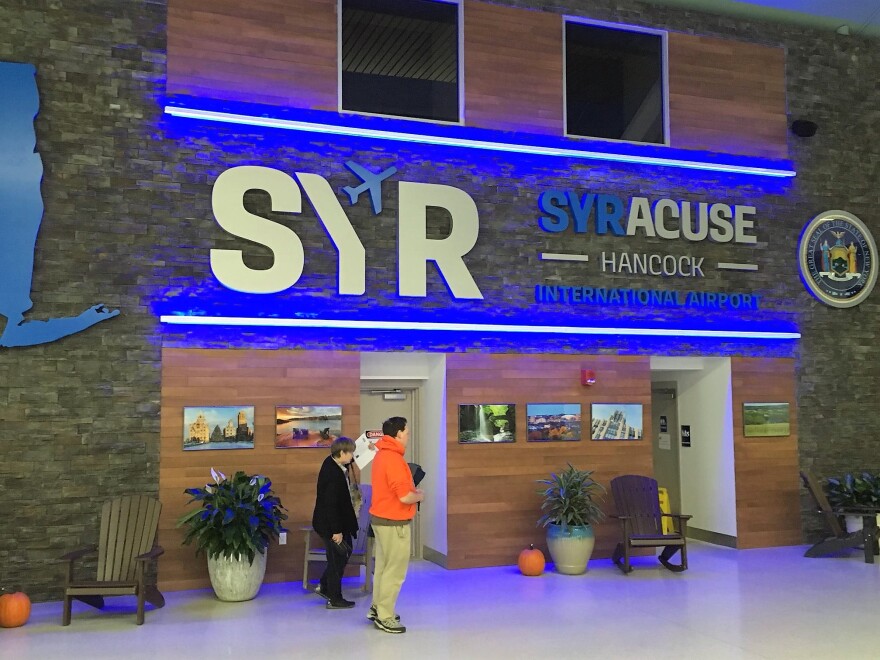 Two people stand in front of a neon and metal sign depicting the logo of Syracuse Hancock International Airport.